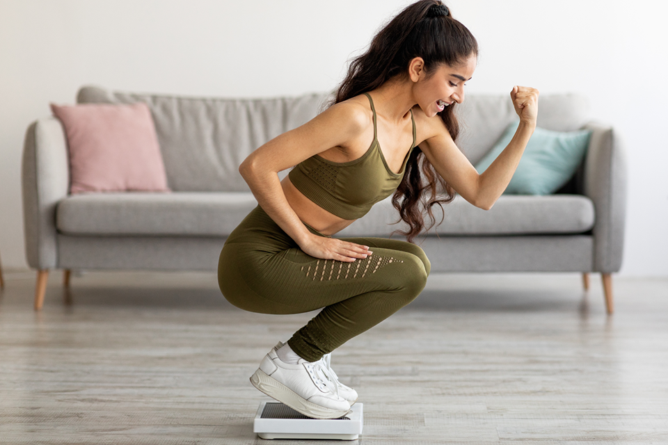 Excited young Indian woman in sportswear sitting on scales, making YES gesture, happy with result of her weight loss success